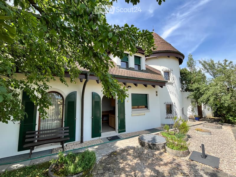 Two-story house with white walls, green shutters, and a red-tiled roof. The house features a round tower with a conical roof and a balcony. The surrounding garden includes a stone bench, potted plants, and a circular stone planter.