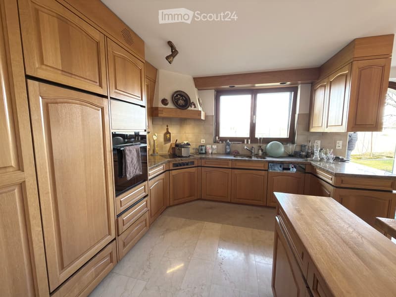 Bright kitchen with light wooden cabinets, granite countertops, and stainless steel appliances including a refrigerator, oven, and dishwasher. The space features a central island and ample natural light from large windows.