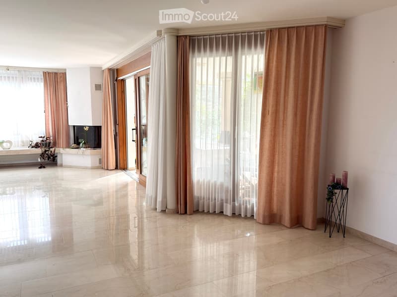 Bright, spacious living room with large windows and sliding glass doors leading to a balcony. Features include a polished marble floor, light-colored walls, and a small side table with candles and a plant. A TV screen is mounted on the wall near the windo