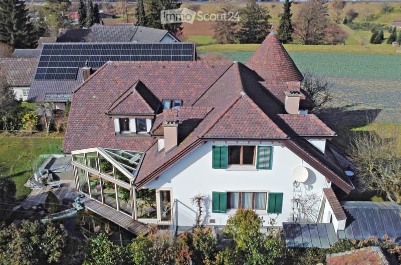 A two-story house with a red-tiled roof, white walls, green shutters, and a large glass sunroom attached to the side. The house features solar panels on the roof and is surrounded by a well-maintained garden and open fields.