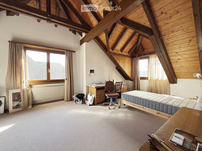 Bright attic bedroom with wooden beams, large windows, a bed, desk, chair, and a small table with magazines.