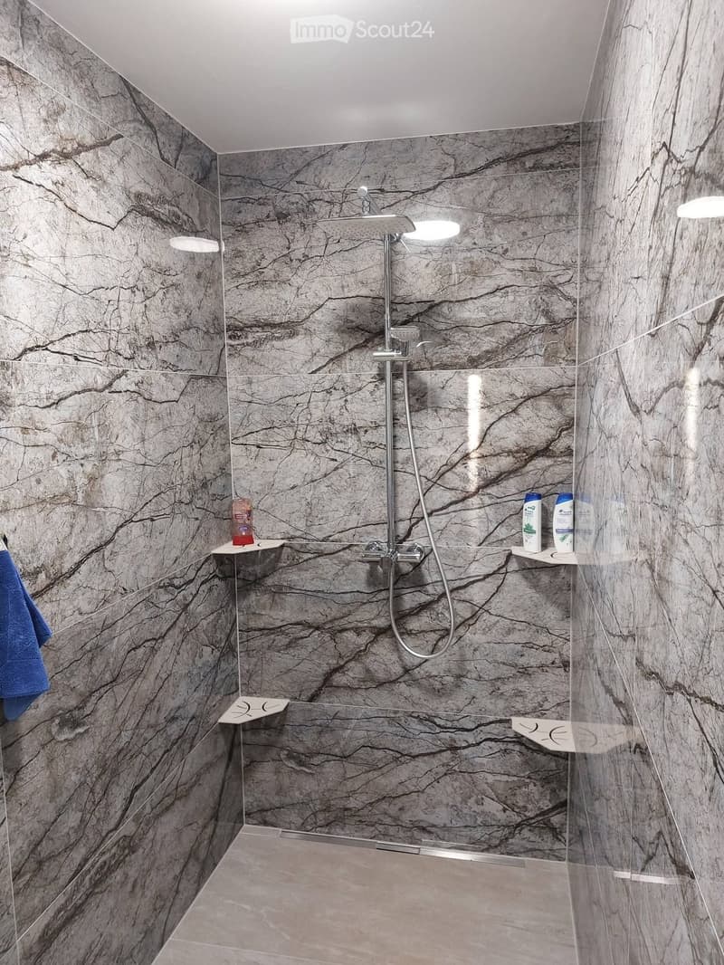 Modern shower area with marble-patterned tiles, glass enclosure, and built-in shelves holding toiletries and a towel.