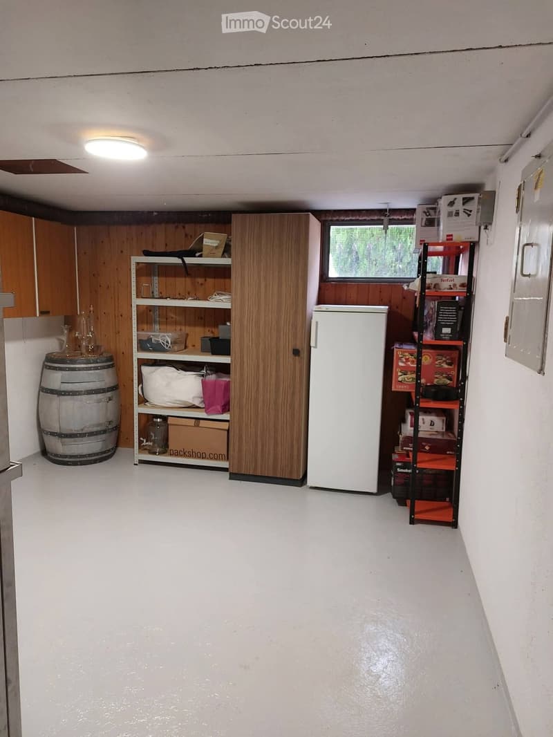 Storage room with white walls, wooden paneling, and a concrete floor. Contains a refrigerator, shelves with various items, a wooden cabinet, and a barrel.