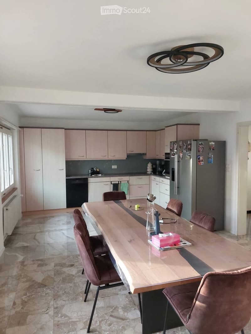Modern kitchen with light-colored cabinets, a long wooden dining table, and brown upholstered chairs. Appliances include a refrigerator and dishwasher.