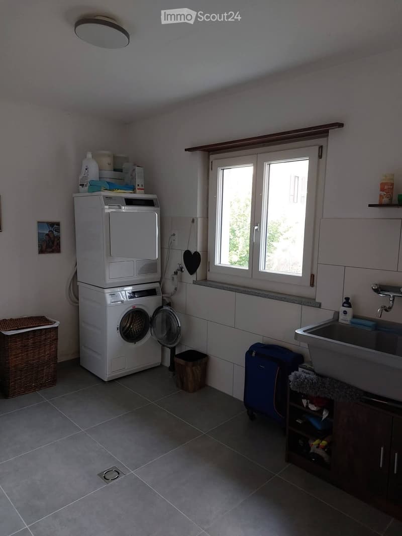 A well-lit laundry room with a washing machine and dryer stacked, a sink with a faucet, a wicker basket, a suitcase, and a small cabinet.