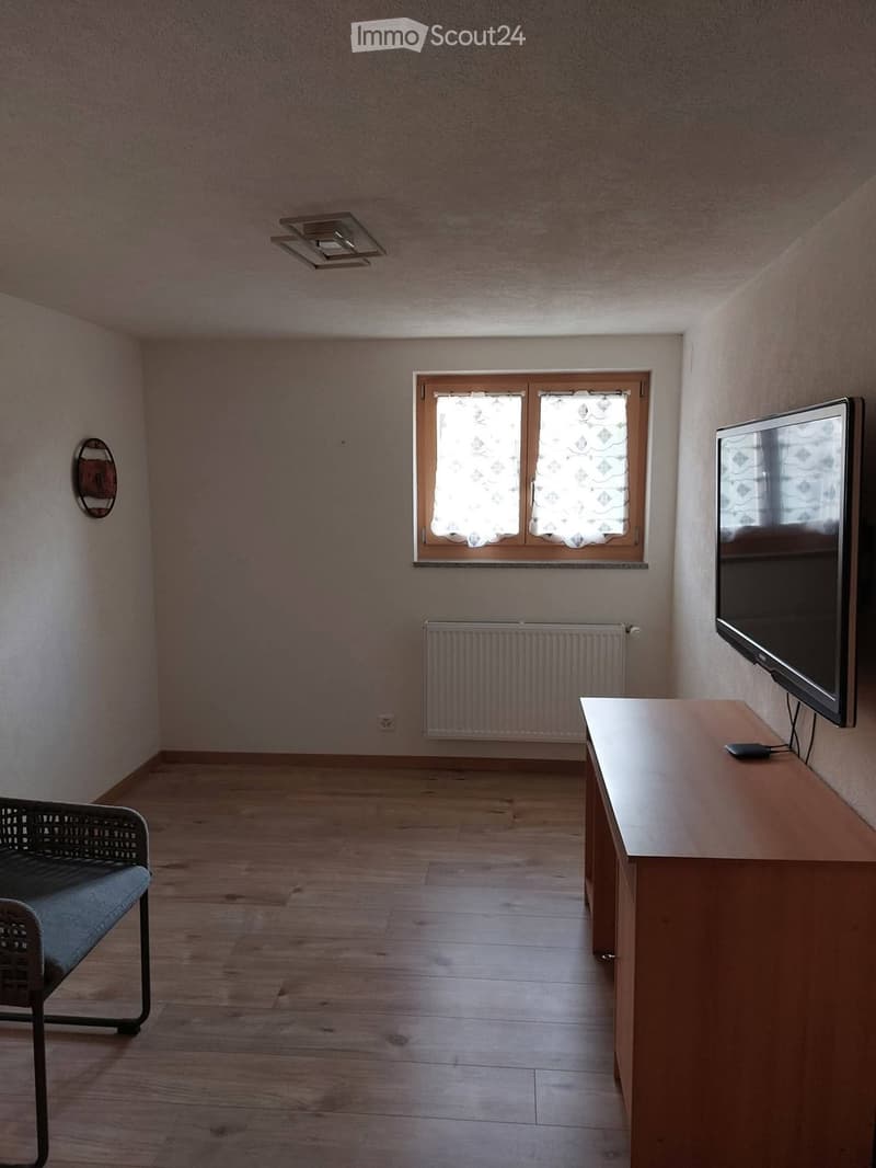 A small, unfurnished room with wooden flooring, a window with curtains, a radiator, a chair, a table, and a television.