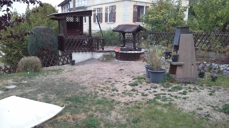 Garden area with a wooden gazebo, a small fountain, potted plants, and a stone barbecue. The yard is bordered by a wooden fence and has a mix of grass and dirt ground.