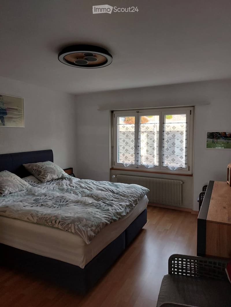 Bedroom with wooden flooring, large window with patterned curtains, bed with patterned duvet, chair, small table, and picture frames on walls.