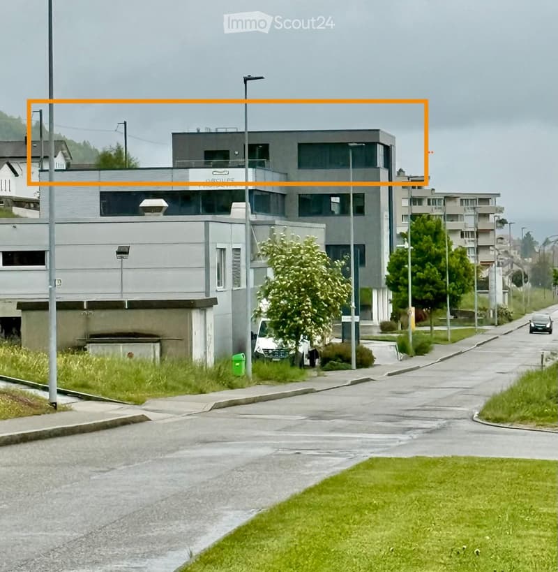Modern multi-story building with a flat roof and large windows, situated on a street with surrounding greenery and a paved road.