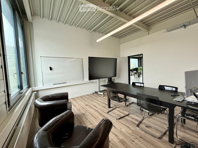 Modern office space with a large conference table, chairs, a sofa, a screen, and a whiteboard. Large windows provide natural light.