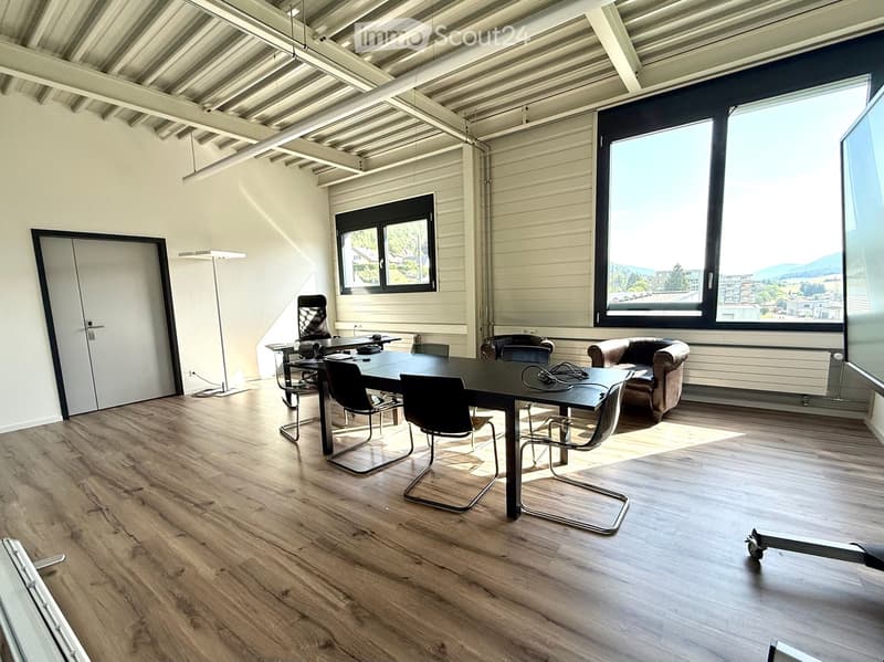 Modern office space with wooden flooring, large windows, a conference table with chairs, a sofa, and a floor lamp.