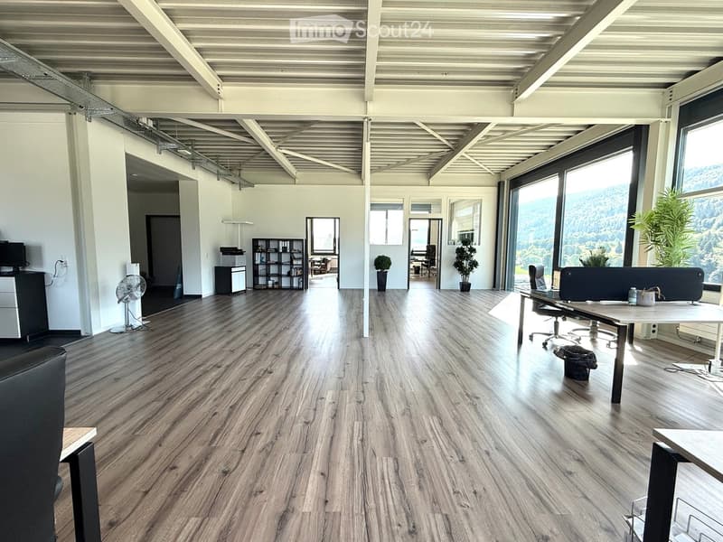 Open-plan office space with wooden flooring, large windows, and modern furniture including tables, chairs, and a fan.