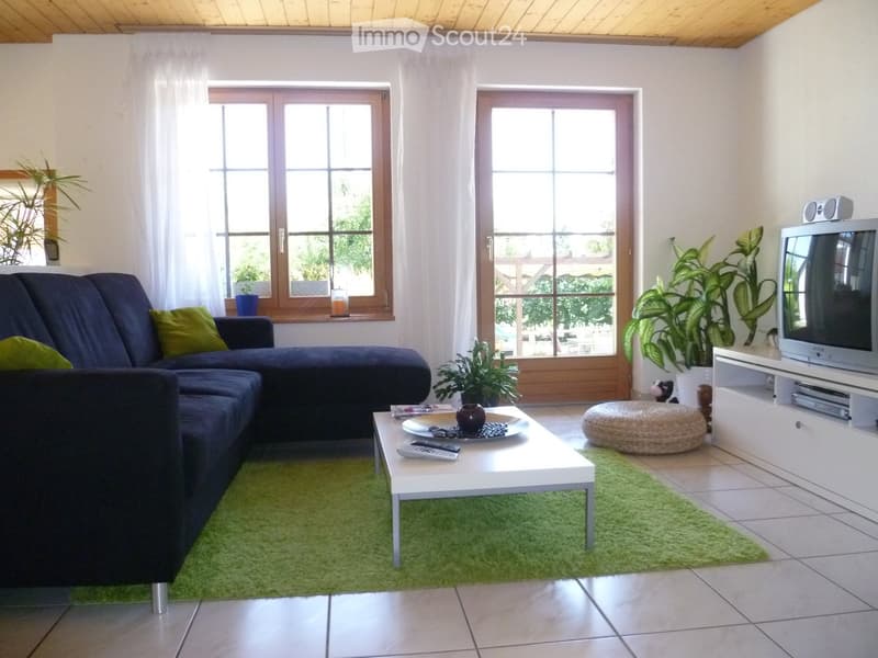 Modern living room with a dark blue sectional sofa, white coffee table, ottoman, TV stand, and plants. Large windows with white curtains and a green rug.