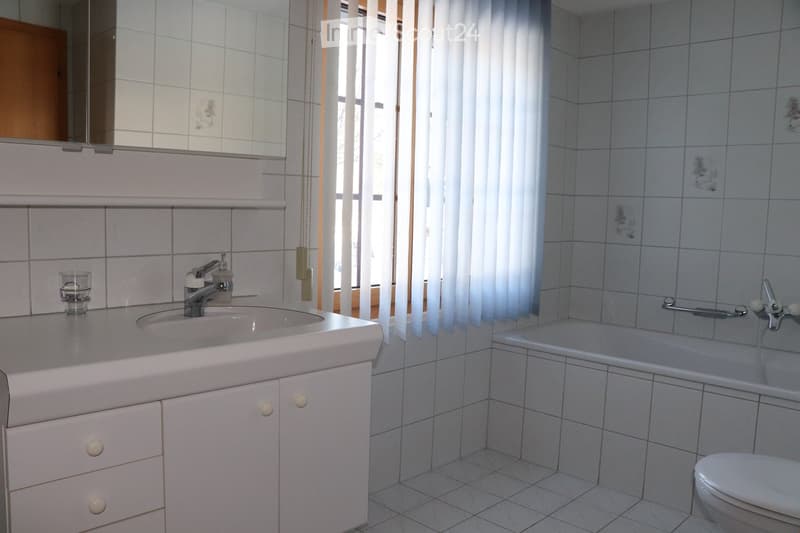 Bathroom with white tiled walls and floor, a sink with a faucet, a bathtub with a handheld shower, a toilet, a mirror above the sink, and a small glass cup on the counter.