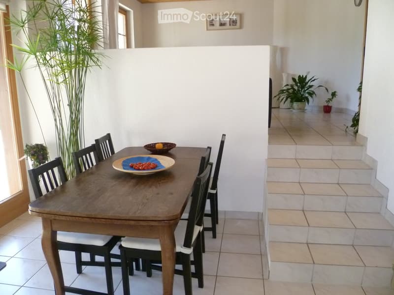 Dining room with a wooden dining table, six chairs, and a bowl of fruit on the table. The room has tiled flooring and large windows with plants visible outside.