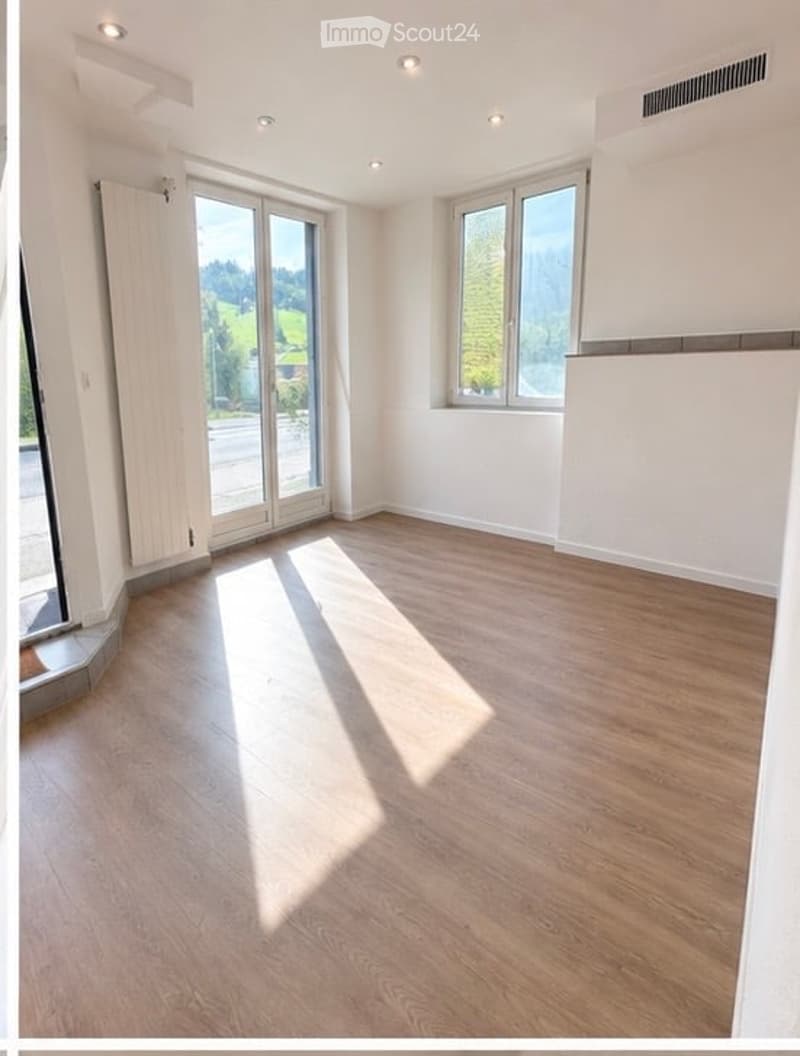 Empty room with light wooden flooring, white walls, large windows, and a sliding glass door leading to a balcony.