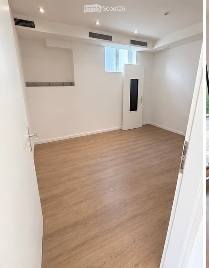 Empty room with wooden flooring, white walls, a single white door, and a small window above the door. No furniture or equipment present.