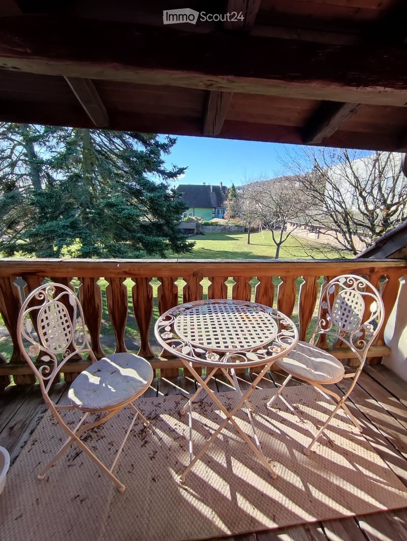 Wooden balcony with a view of a green landscape, featuring a small round table and two matching chairs.