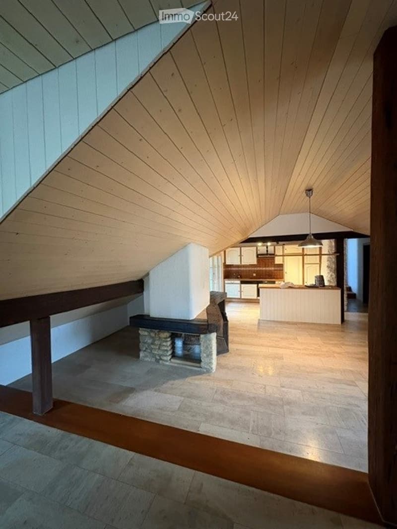 Loft-style living space with exposed wooden beams, a stone fireplace, a sofa, and an open kitchen area with white cabinets and a kitchen island.