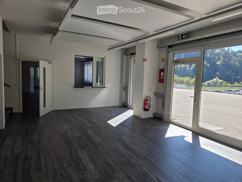 Empty hallway with wooden flooring, white walls, and large windows allowing natural light. Features include a fire extinguisher, an emergency exit sign, and a door leading to another area.
