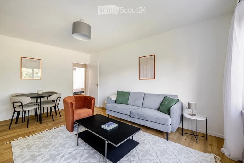 Modern living room with a grey sofa, black coffee table, orange armchair, dining area with a black table and chairs, side table with lamp, and two framed mirrors on the walls.