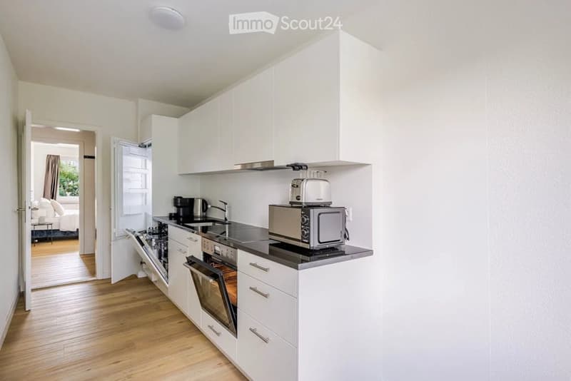 Modern kitchen with white cabinets, stainless steel appliances, and a black countertop. Visible appliances include a refrigerator, dishwasher, oven, microwave, toaster, and coffee maker. The kitchen features a sink with a faucet and is connected to a bedr