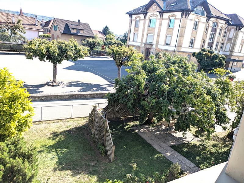 Garden area with grass, trees, a wooden bench, and a paved pathway, with a parking lot and residential buildings in the background.