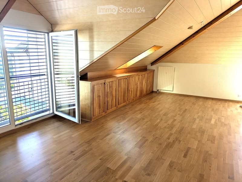 Bright, empty room with wooden flooring, built-in wooden cabinetry, large windows with blinds, and a slanted ceiling with exposed wooden beams.