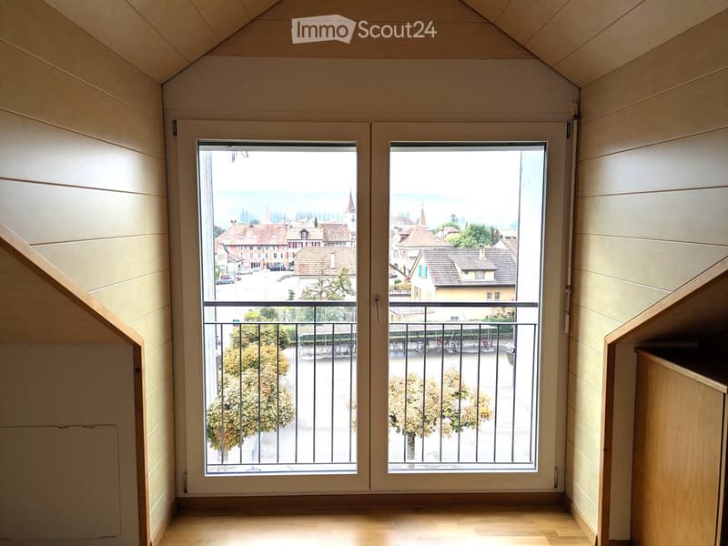 Balcony with wooden floor, large windows, metal railing, overlooking a town with visible buildings and trees.