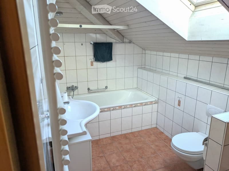 Bathroom with white tiles, a bathtub, a sink, a toilet, and a towel hanging on a rack. The room features a slanted wooden ceiling and a small window at the top.