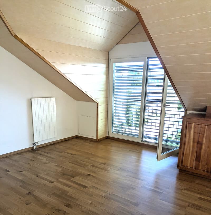 Empty attic room with wooden flooring, white walls, and a large window with blinds. A radiator is visible on one wall.