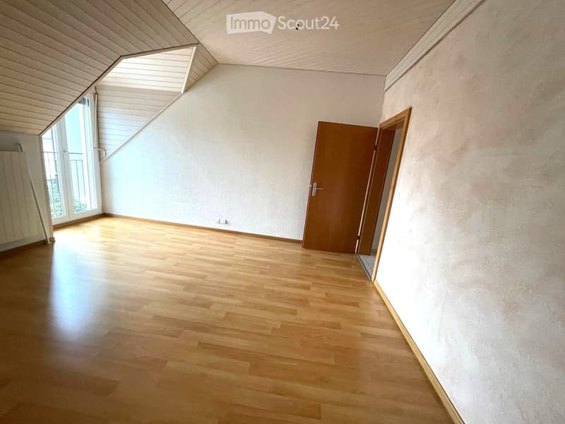 Empty room with wooden flooring, white walls, and a large window with blinds. There is a wooden door and a slanted ceiling with a skylight.