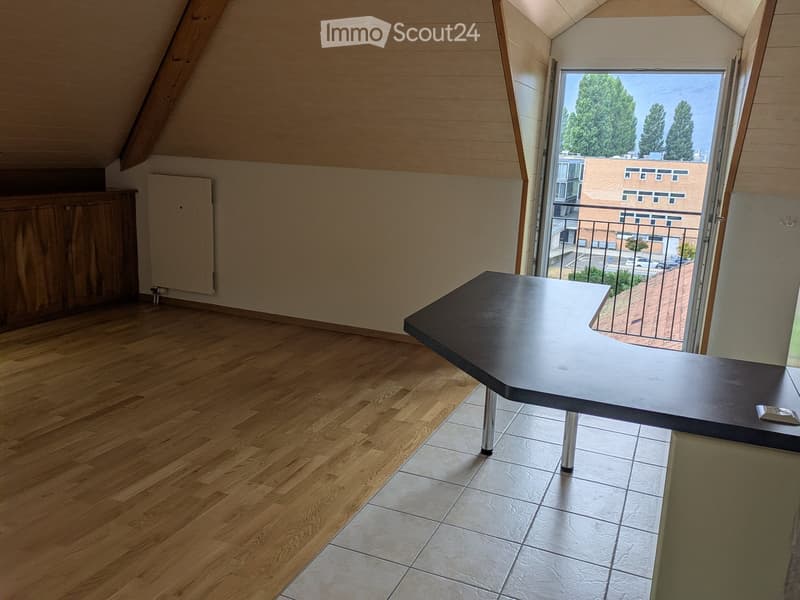 Bright, airy attic room with wooden flooring, large window offering a view, a sleek black table with metallic legs, and a wooden cabinet.