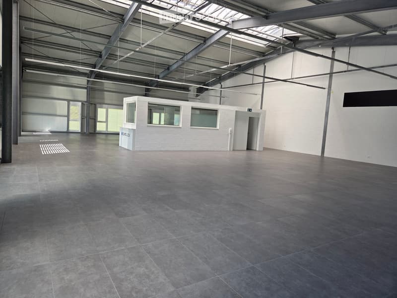 Large, empty industrial-style room with concrete floors, exposed metal beams, and large windows. A small white booth with a door and windows is visible in the center.