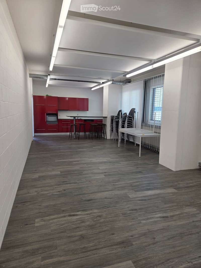 Open-plan room with red fitted kitchen cabinets, an oven, a central table with stools, and a stack of chairs near a window with blinds.