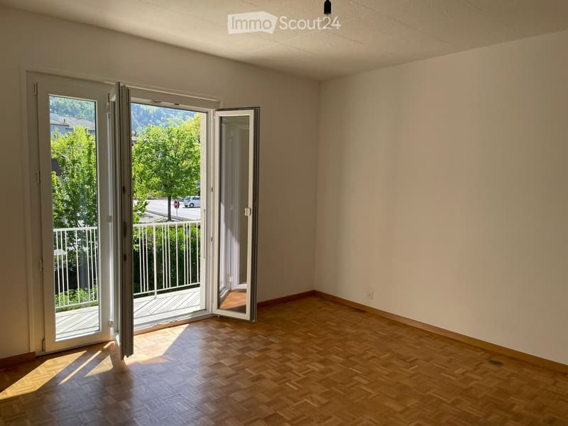 Empty room with wooden flooring, white walls, and large sliding glass doors leading to a balcony with a view of greenery and a street.