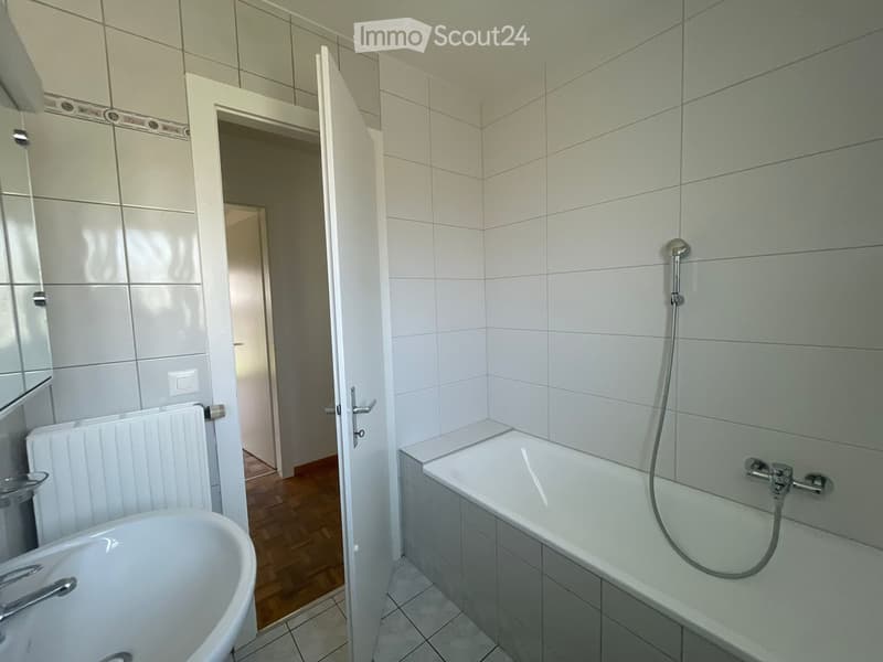 Modern bathroom with white tiled walls, a bathtub with a handheld shower, a sink, and a radiator. The door to the adjacent room is open, revealing a wooden floor.
