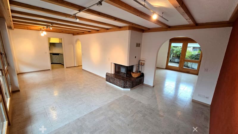 Bright, open living space with exposed wooden beams, tiled floor, fireplace, and large windows leading to an outdoor area.