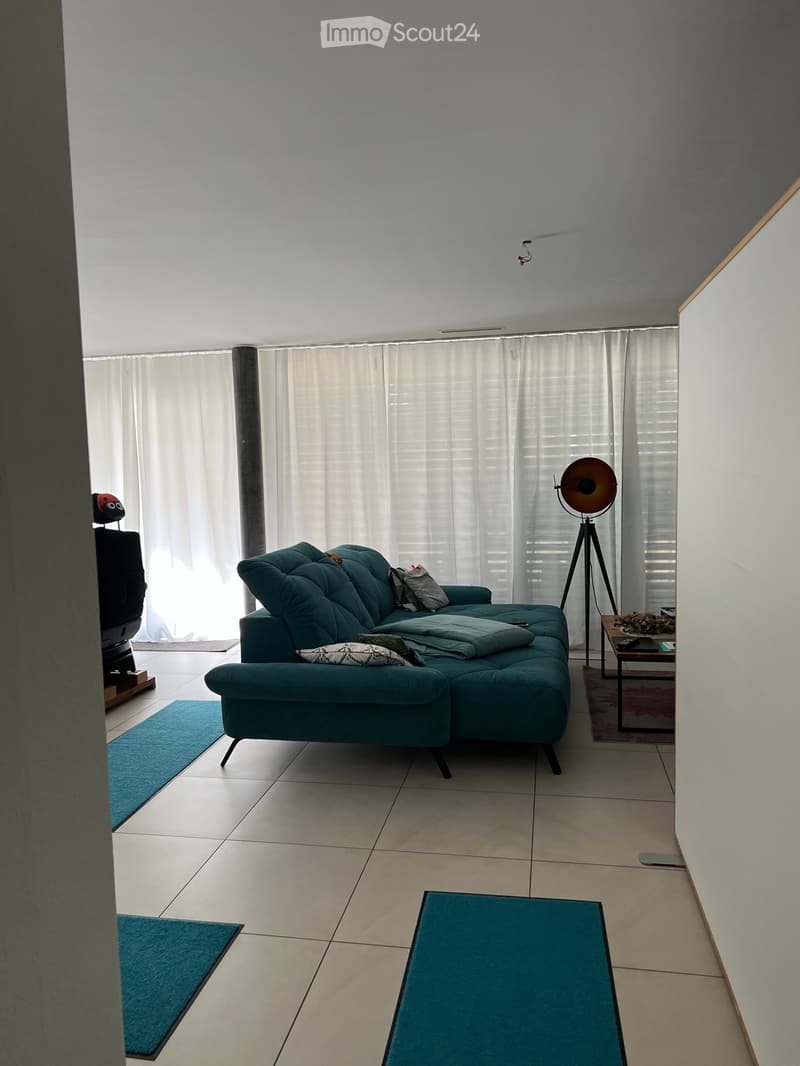 Modern living room with a teal sofa, a small wooden side table, a floor lamp, and a television. The room features large windows with white curtains and tiled flooring.