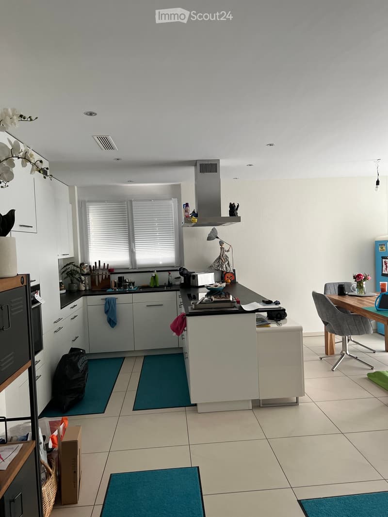 Modern kitchen with white cabinets, a central island, and a dining area. Features include a sink, stove, refrigerator, and various kitchen items on the countertops.