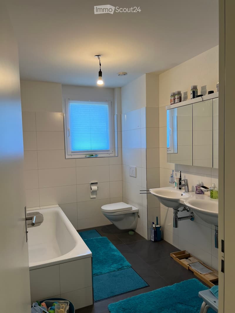 Modern bathroom with a bathtub, toilet, double sink, mirror, and various toiletries. Features tiled walls, a window with blinds, and blue bath mats.