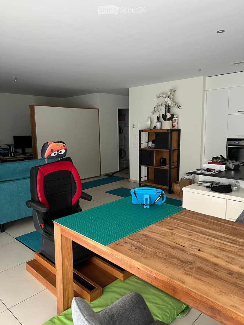 A modern, well-lit room with a wooden table, ergonomic chair, shelf with books and decorative items, sofa, and a small kitchen area with an oven and countertop.