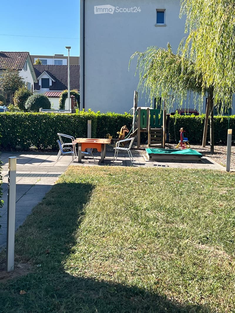 Garden area with a small seating area featuring a table and chairs, a playground with swings and a slide, and a grassy lawn.