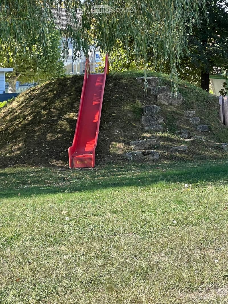 A grassy yard with a red plastic slide positioned on a dirt mound, surrounded by trees and a small structure in the background.