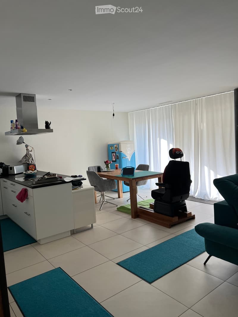 Open-plan living and dining area with a modern kitchen island, a dining table with chairs, a sofa, and a refrigerator. The space features large windows with curtains and tiled flooring.