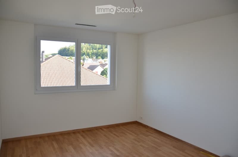 Empty room with wooden flooring and white walls, featuring a large window with a view of neighboring rooftops and greenery.