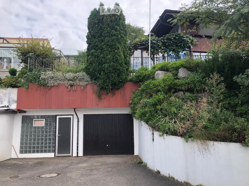 Two-story building with a red roof, white walls, and a black garage door. The building has a landscaped rooftop with greenery and a metal railing.