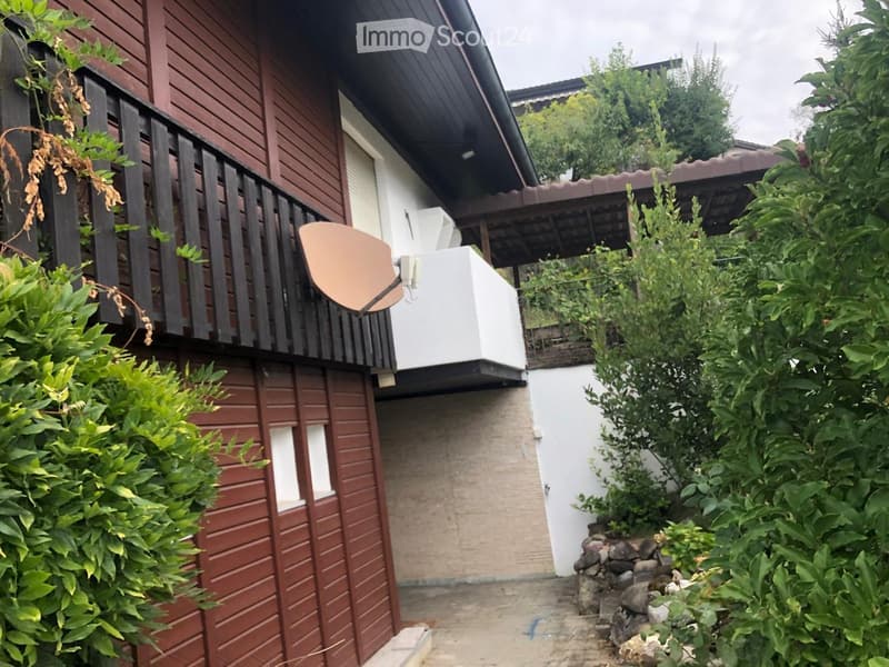 Two-story house with wooden exterior, balcony, and satellite dish. Surrounded by greenery and a stone pathway.