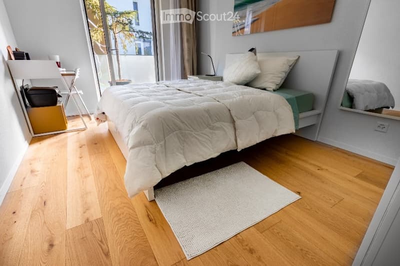 Cozy bedroom with wooden flooring, large window with view, bed with white bedding, nightstand, mirror, small chair, and table.