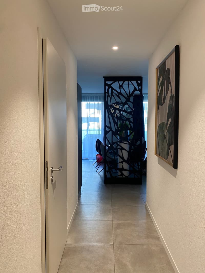 Clean, modern hallway with tiled floor, white walls, and a decorative black screen at the end. A framed picture hangs on the right wall, and a door is visible on the left.